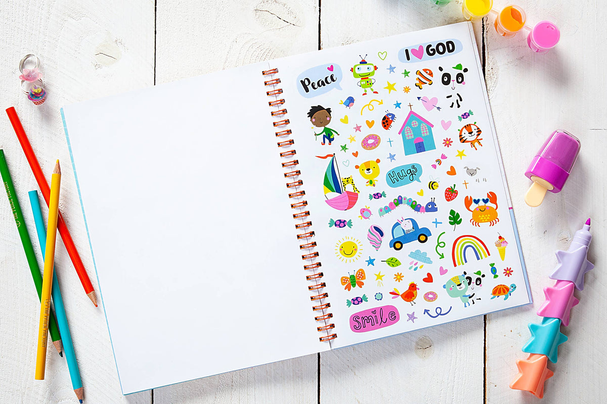 The “God Made Me Journal Book” by BroadStreet Publishing Group, LLC is open on a white wooden table, displaying colorful stickers on one page and surrounded by colored pencils, a pink marker, a small bottle, and star-shaped crayons.