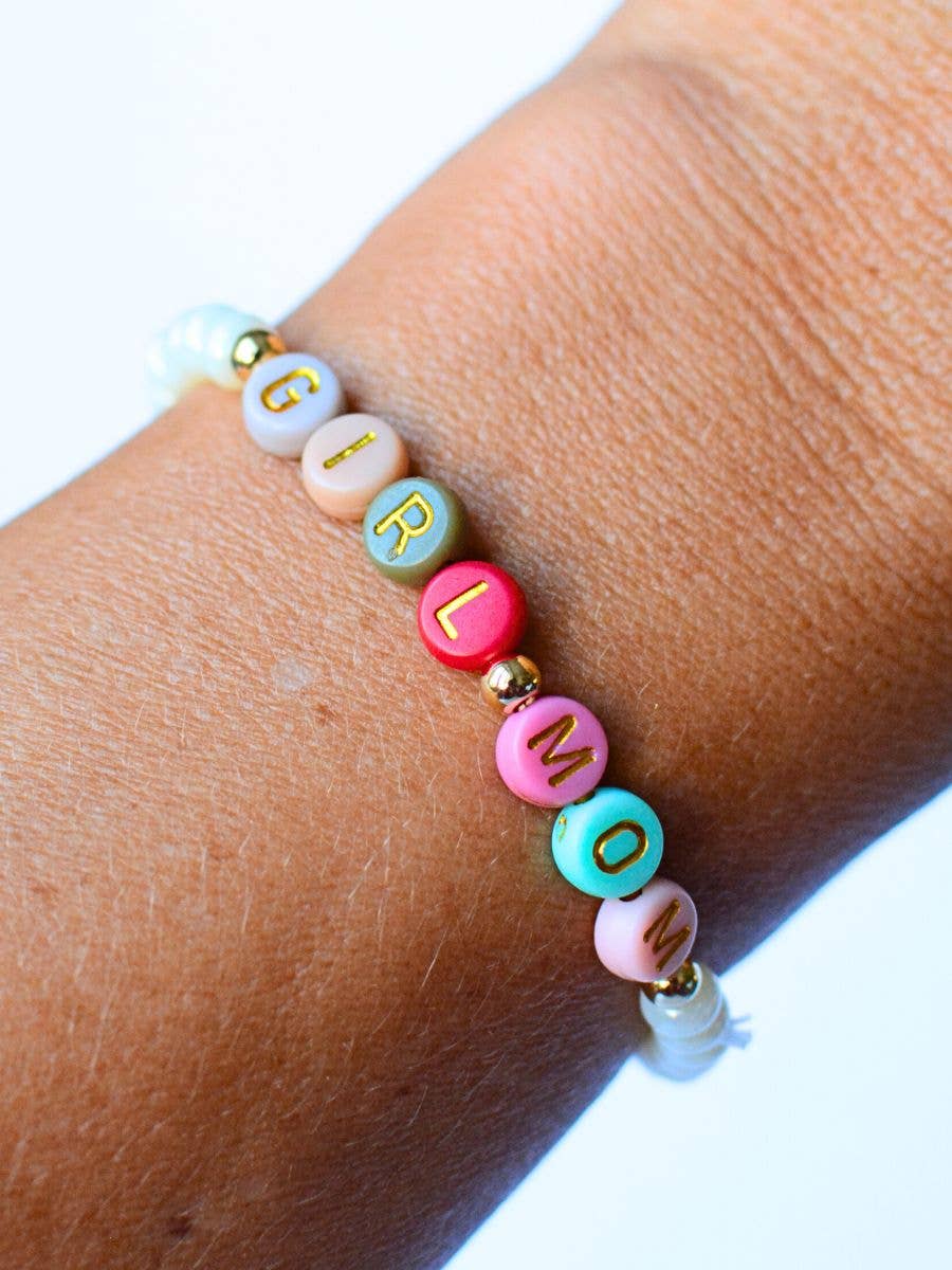 A close-up of a wrist adorned with the Girl Mom Beaded Bracelet by Two and Crew - Faith Based Jewelry & Gifts. The bracelet features colorful round beads in pink, red, green, white, and gold, with gold letters spelling “GIRL MOM,” set against a light blue background.