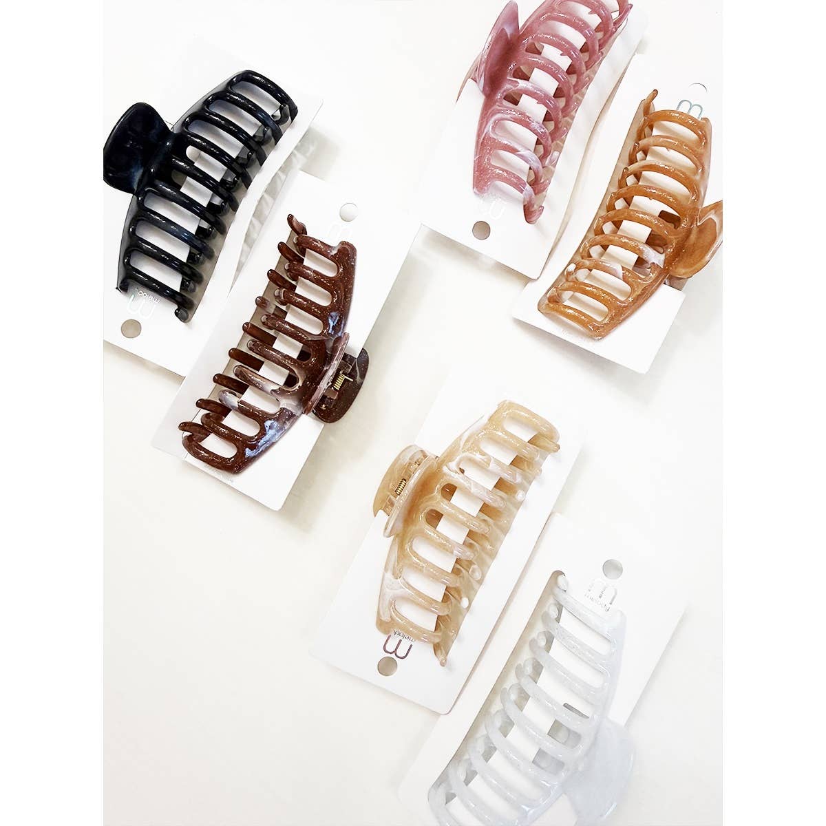 Five Marbled Round Comb Hair Claw Clips from Love and Repeat—featuring large sizes in black, brown, pink, beige, and white—are displayed individually on white cards against a light background, arranged in a scattered pattern.