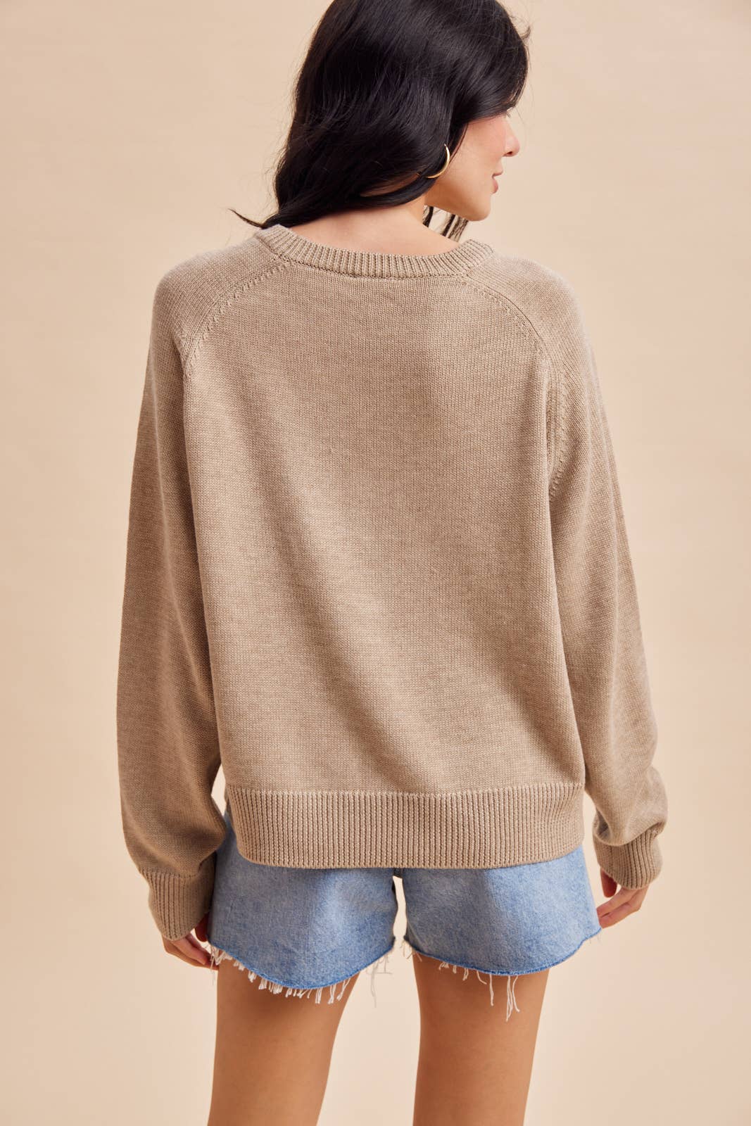 A person seen from the back, wearing a tan, long-sleeved Mallard Duck Knitted Sweater by In Loom and light-wash denim shorts.