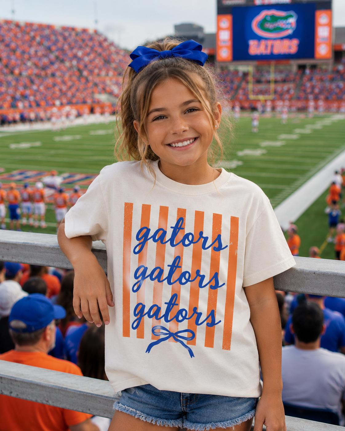 A young girl wearing a blue bow and a Gators Stripe Graphic t-shirt from Three Threads Branding Co. smiles at a football stadium, with fans and the game visible in the background.