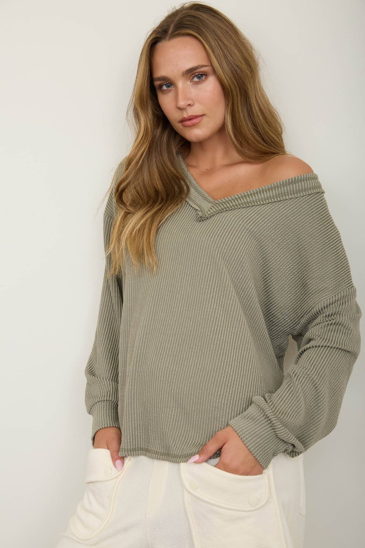 A woman wearing the Blu Pepper Olive V-Neck Long Sleeve Top and white pants stands against a plain background, looking at the camera with a relaxed expression and one hand in her pocket.
