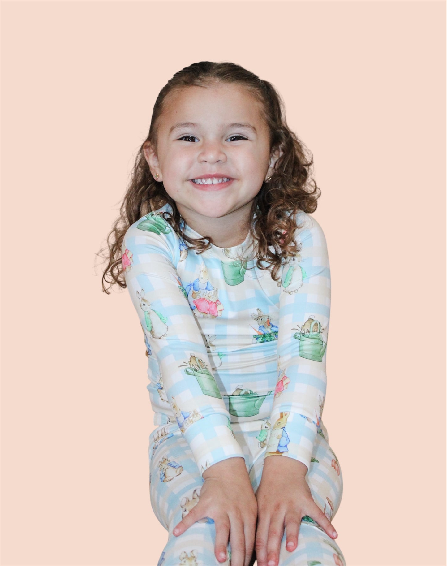 A smiling young child in the Peter's Easter Picnic Jammies Set by Tiny Tot Bundles sits against a pale peach background, hands resting in front and looking at the camera.