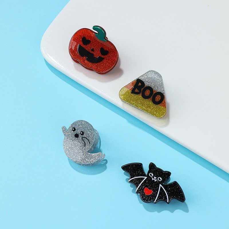 Four glittery Halloween-themed mini hair clips from JOSSLYN by wall to wall—a smiling pumpkin, a candy corn with “BOO” written on it, a waving ghost, and a bat with a red heart—arranged on a white and blue surface.