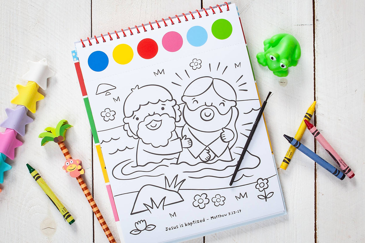 A page from the Cozy Club Bible Friends Art Pad Book by BroadStreet Publishing Group, LLC features a children’s coloring scene of Jesus being baptized, surrounded by crayons, a paintbrush, colorful erasers, and a green frog toy on a white wooden surface. The coloring page is open on its spiral-bound pad.