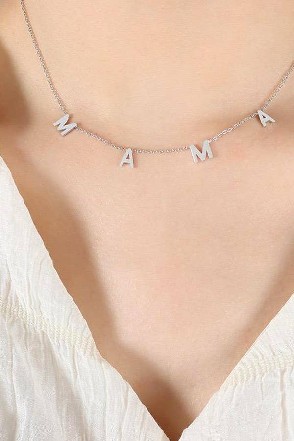 A close-up of the Mama Tarnish Free Necklace by JOSSLYN by wall to wall, featuring hanging letter charms that spell “MAMA” in silver, worn around a person’s neck with a white v-neck top.