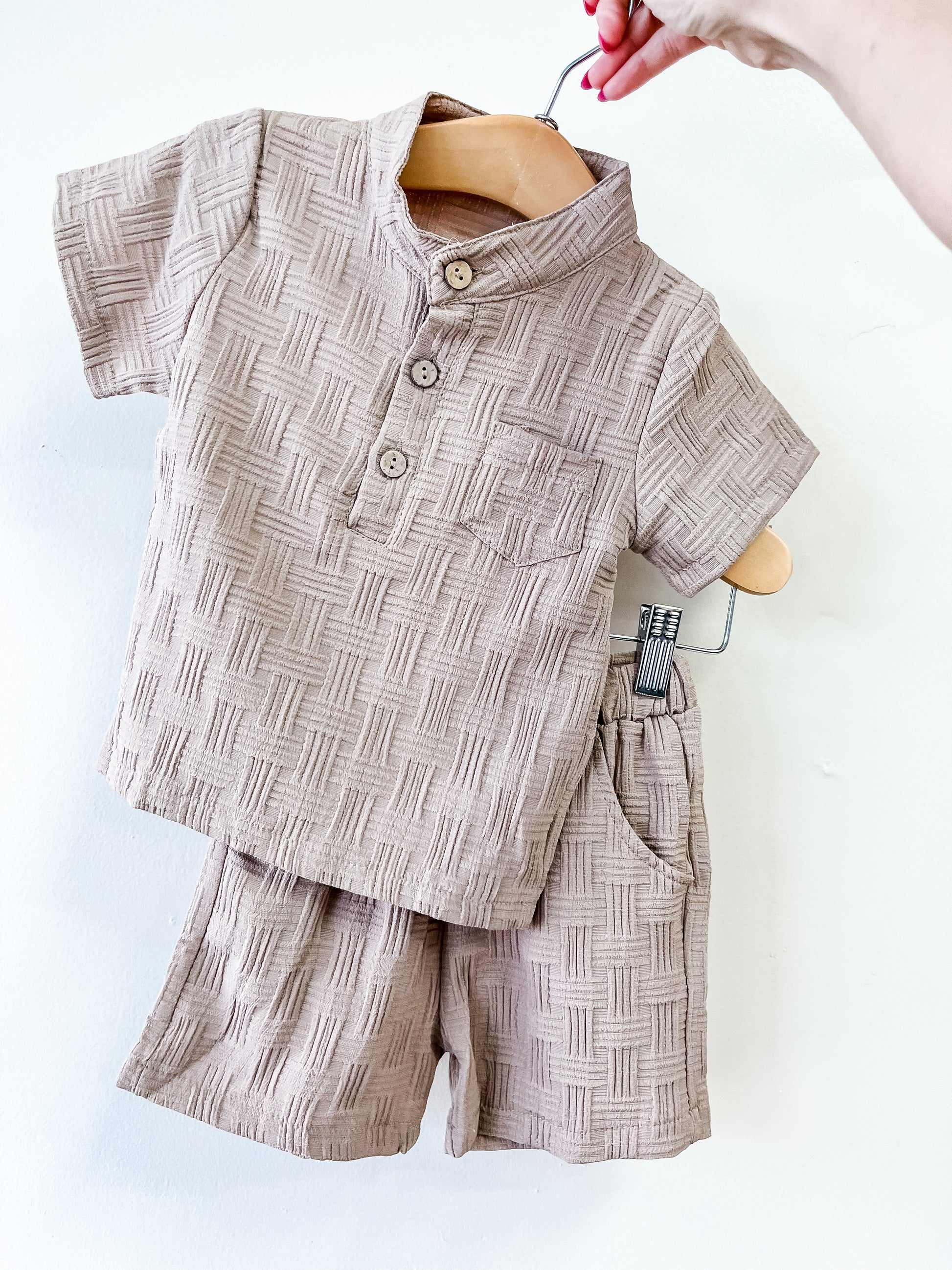 The Tiny Tot Bundles Textured Boy Button Up Set, featuring a beige short-sleeve shirt with three buttons and matching shorts, hangs on a wooden hanger held by hand against a plain white background.