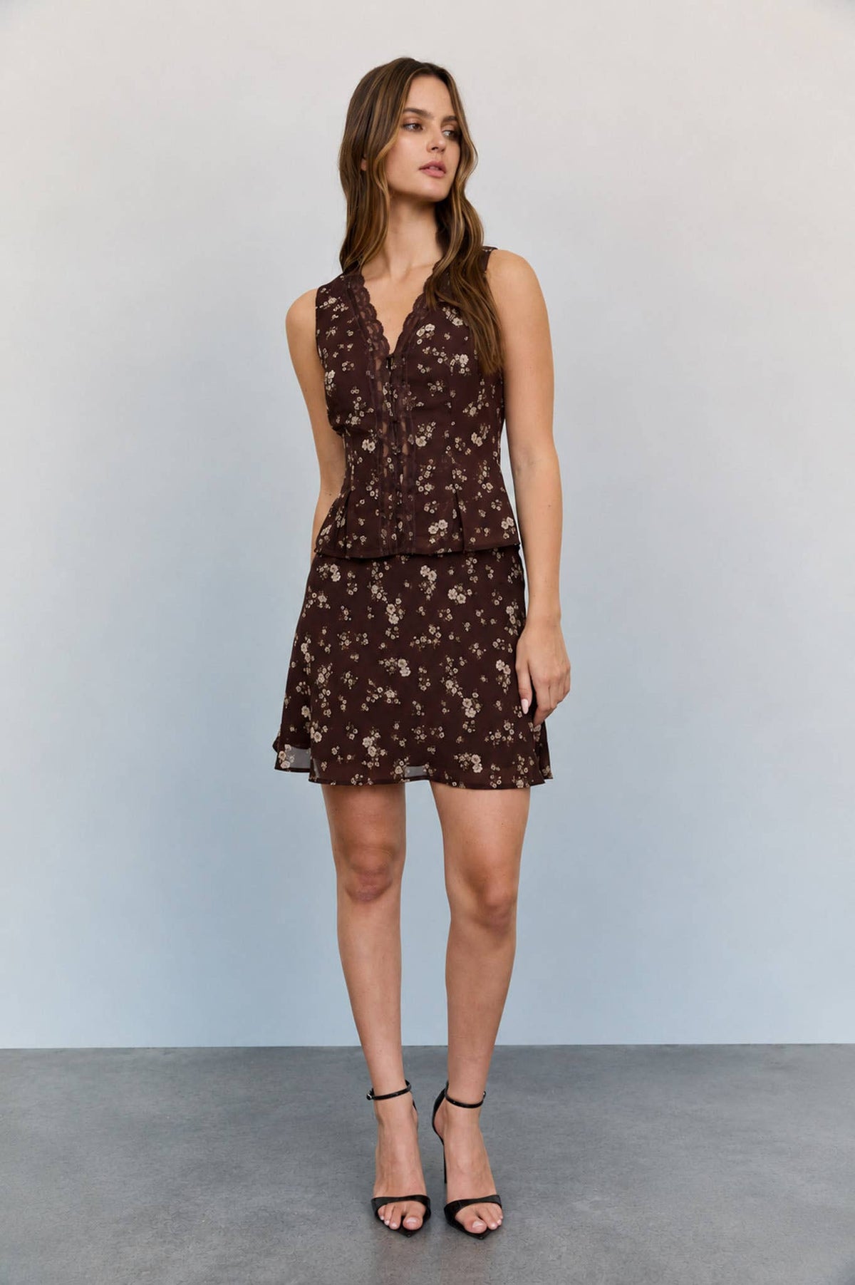 A woman stands against a plain background, wearing The Workshop Studio’s Floral Lace Blouse and black high-heeled sandals. She has long hair, looks slightly to the side, and rests one hand at her side.
