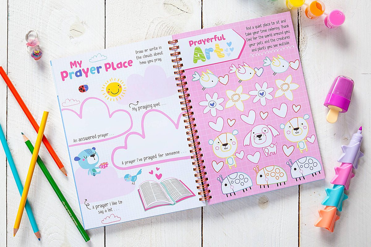 The God Made Me Journal Book by BroadStreet Publishing Group, LLC is open on a white wooden surface, featuring colorful illustrations of birds, clouds, hearts, flowers, a dog, and ladybugs. Pencils, tiny bottles, and star-shaped crayons are placed beside it.