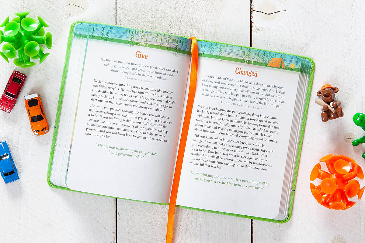 The A Daily Word for Boys Book by BroadStreet Publishing Group, LLC lies open with a ribbon bookmark on a white wooden surface, surrounded by small toy cars, green and orange sensory toys, and a brown teddy bear.