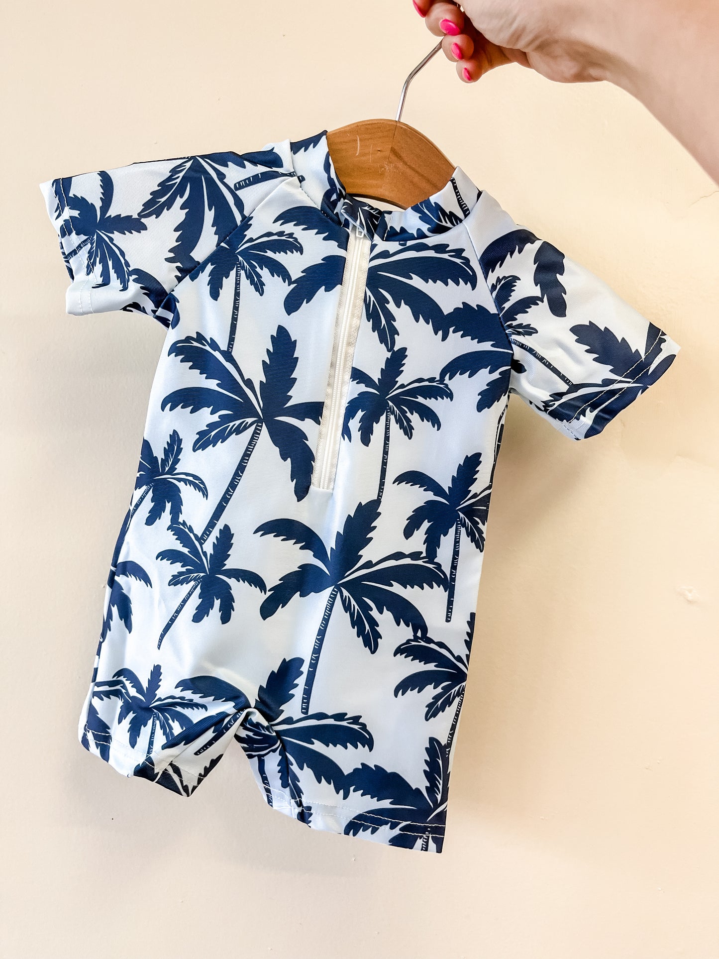 A hand holds a Palm Coast Swimsuit by Tiny Tot Bundles, featuring a navy blue palm tree print on white, displayed on a wooden hanger against a light beige wall.