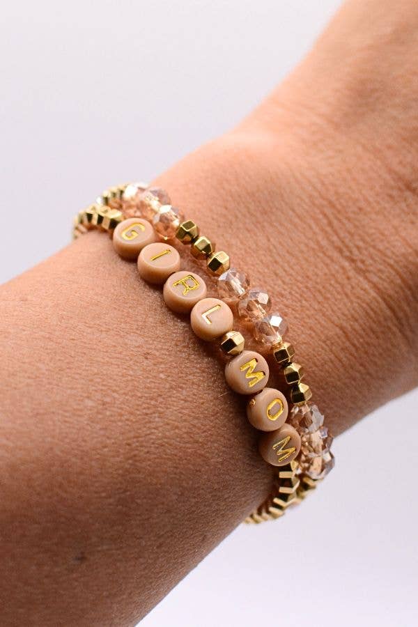 A close-up of a wrist wearing the Girl Mom Bracelet Set by Two and Crew - Faith Based Jewelry & Gifts, featuring three bracelets: one with round beige beads spelling GIRL MOM in yellow letters, and two accented with gold and clear beads.