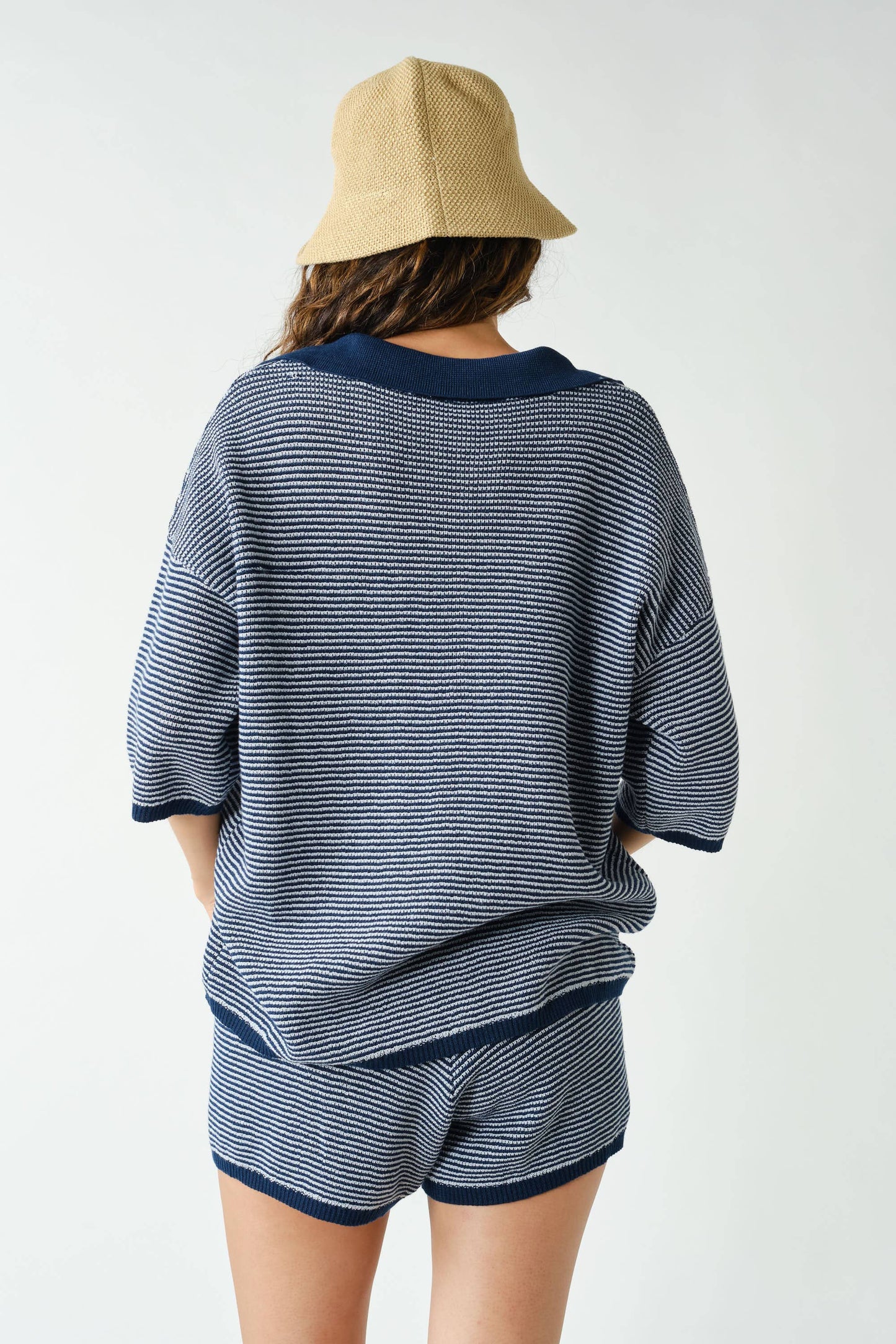 A person wearing the Mono B Navy Coastal Breeze Knit Set—a blue-and-white striped top with matching shorts—and a beige hat stands facing away from the camera against a plain light background.