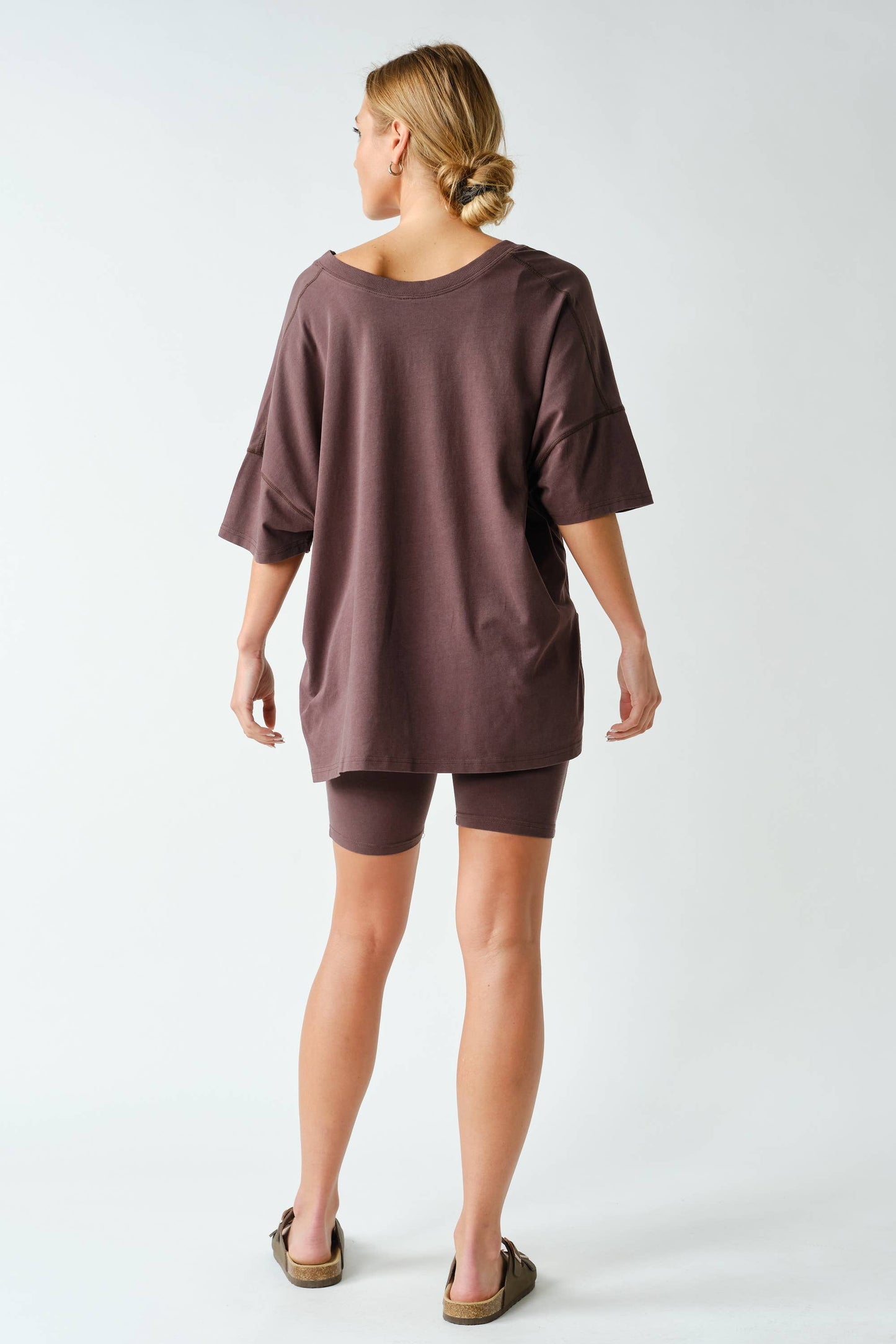 A woman stands facing away, wearing the Mono B Switchback Pima Cotton Set—an oversized brown T-shirt paired with matching fitted shorts—and slip-on sandals. Her hair is tied back in a low bun. The background is plain white.