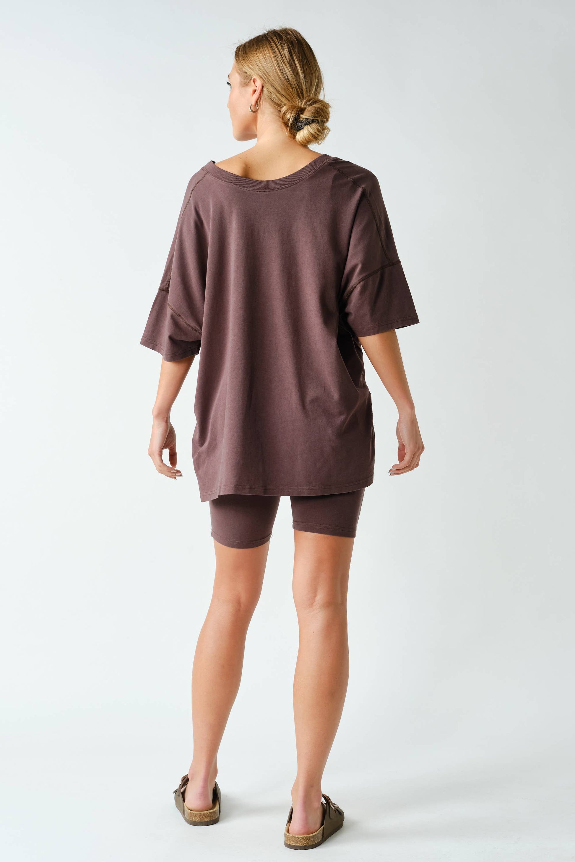 A woman stands facing away, wearing the Mono B Switchback Pima Cotton Set—an oversized brown T-shirt paired with matching fitted shorts—and slip-on sandals. Her hair is tied back in a low bun. The background is plain white.