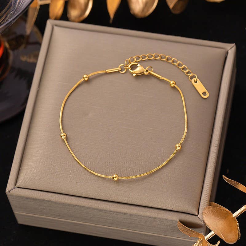 The JOSSLYN by wall to wall Dainty Gold Bracelet, featuring small beads and an adjustable chain, is showcased on a taupe jewelry box and accented by golden decorative leaves.