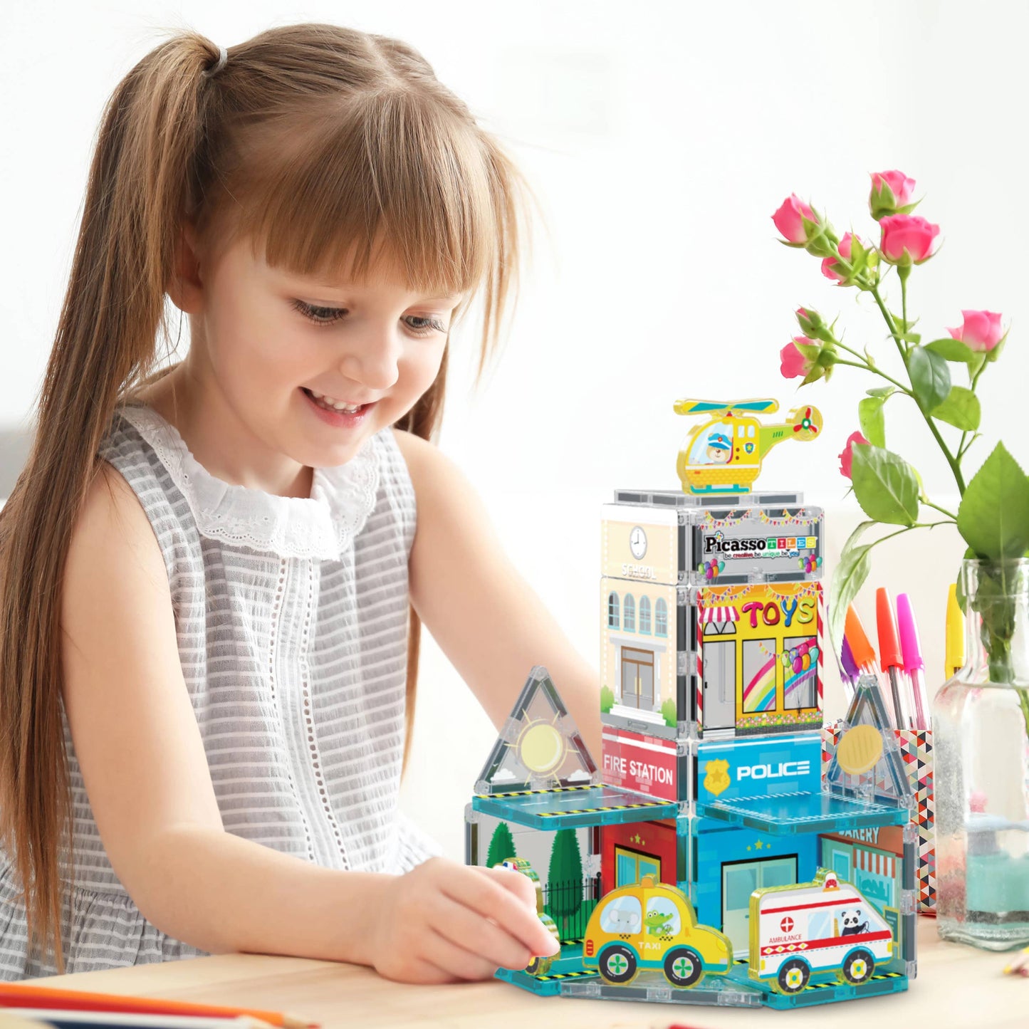 A young girl with pigtails smiles as she plays with the PicassoTiles 26pc City Theme Magnetic Tile Building Set by PicassoTiles, creating buildings, a police car, ambulance, and helicopter on a table with markers and a flower vase.