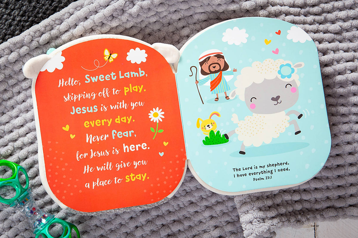 The Squish and Snugg Lamb Book by BroadStreet Publishing Group, LLC is open, displaying one page with colorful flowers and clouds alongside text, while the opposite page features charming illustrations of a shepherd, a joyful lamb, a bunny, and Psalm 23:1 about the Lord as our shepherd; a pacifier rests beside the book.