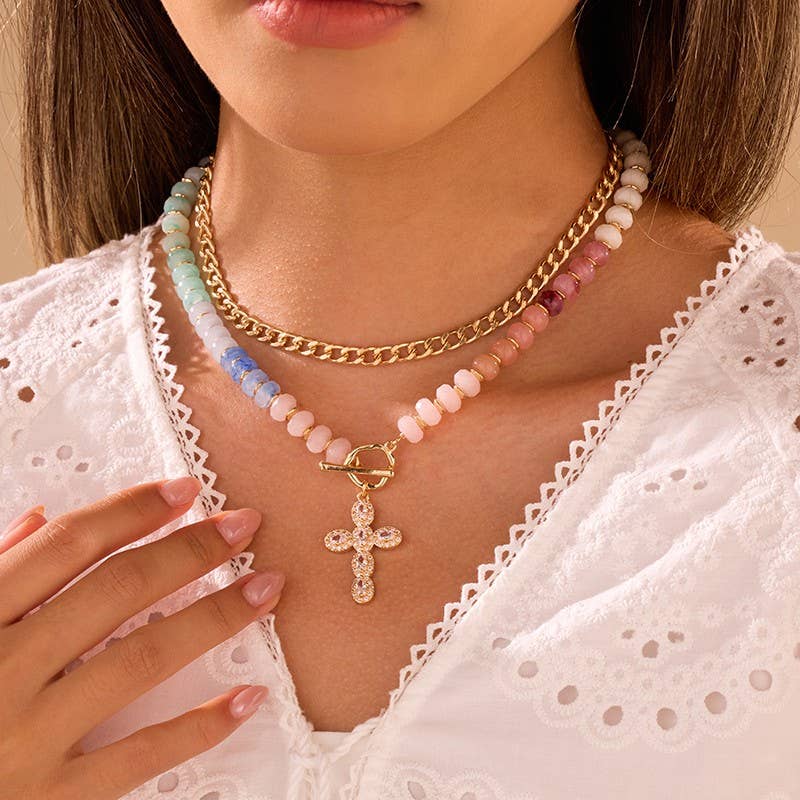A woman wearing a white eyelet blouse showcases the JOSSLYN by wall to wall Multi Layer Pendant Resin Beaded Necklaces, featuring a gold chain, a pastel beaded strand, and a beaded necklace with a crystal cross pendant. Her hand rests gently on her collarbone.