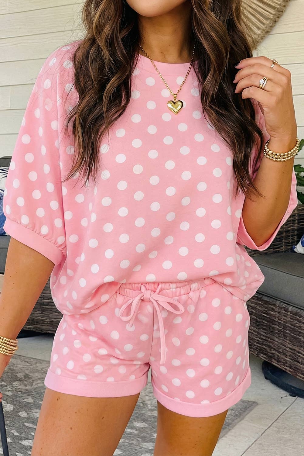 A woman stands indoors wearing the EVELA INC Pink Polka Dot Short Set, which features coordinating shorts and a matching top. She accessorizes with gold bracelets and a heart necklace, and her long brown hair falls in loose waves.