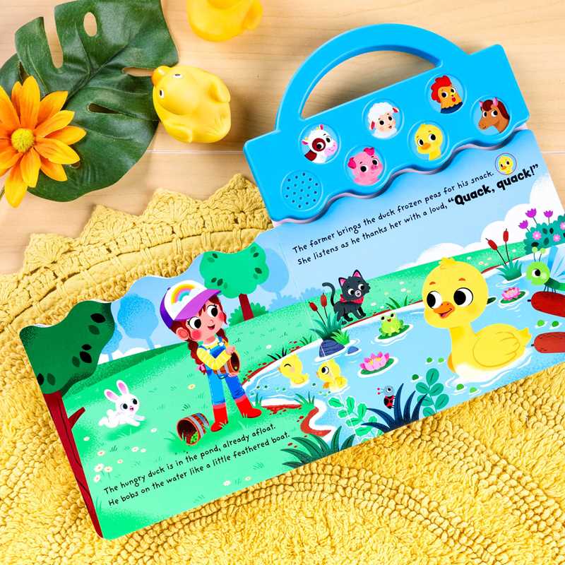 The Moo, Oink, Baa! Book by Simon & Schuster is open to a farm scene with a pond, ducks, a girl, and animals. Nearby are a yellow flower and rubber duck. The book features animal sound buttons on its blue handle.