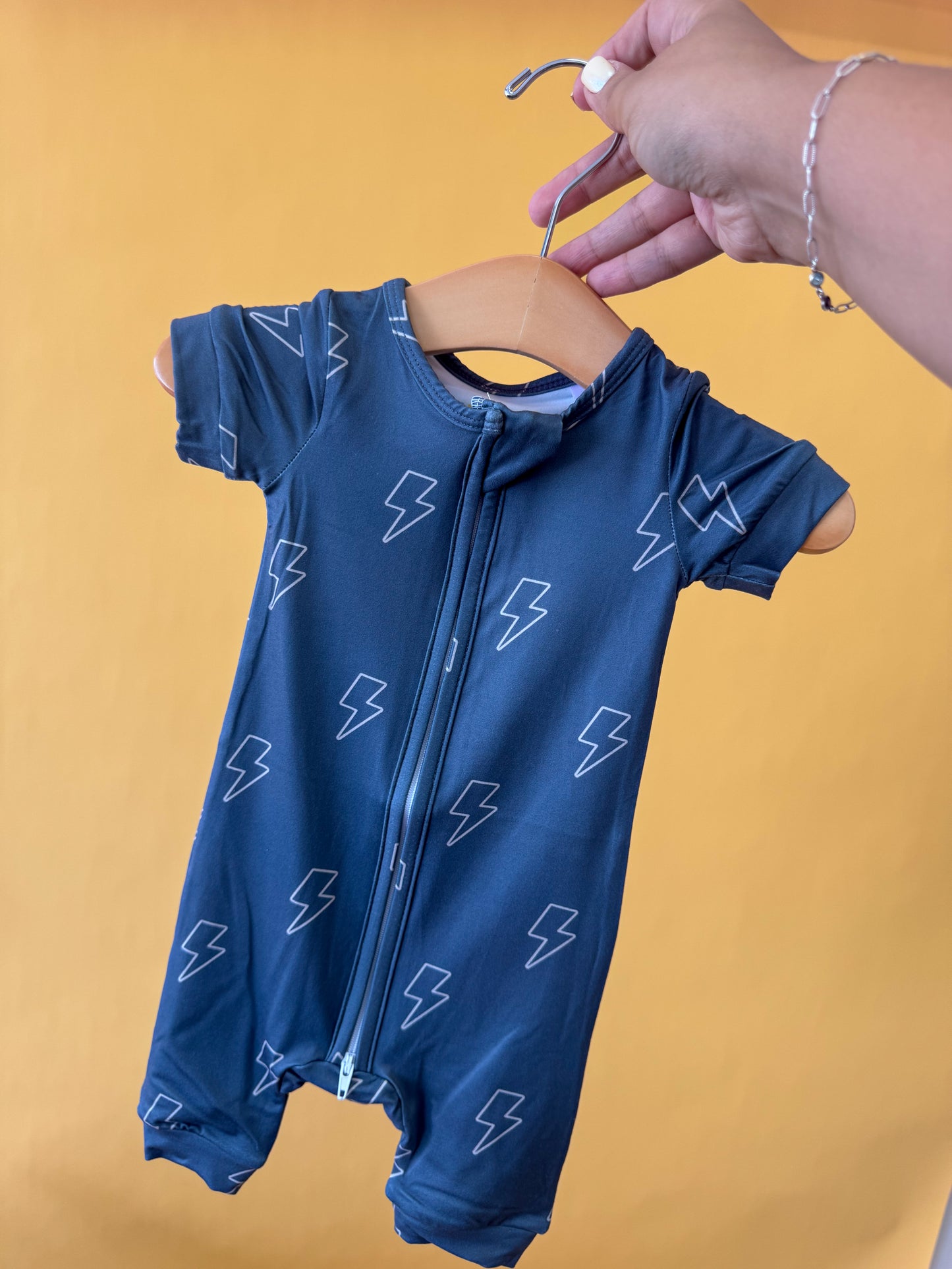 A hand holds a hanger with the Tiny Tot Bundles Storm Chaser Short Zippy, a blue baby romper featuring a front zipper and lightning bolt pattern, against a yellow background.