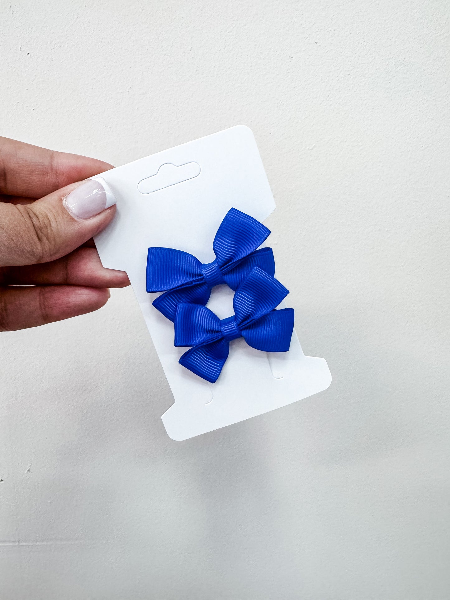 A hand with light nail polish holds a white card displaying two identical blue Duo Piggy Clip Pack hair bows by Tiny Tot Bundles against a plain light background.