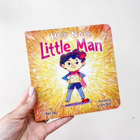 A hand holds the Simon & Schuster “Hey, Now, Little Man Book,” featuring a confident boy with hands on hips on a vibrant, colorful cover.