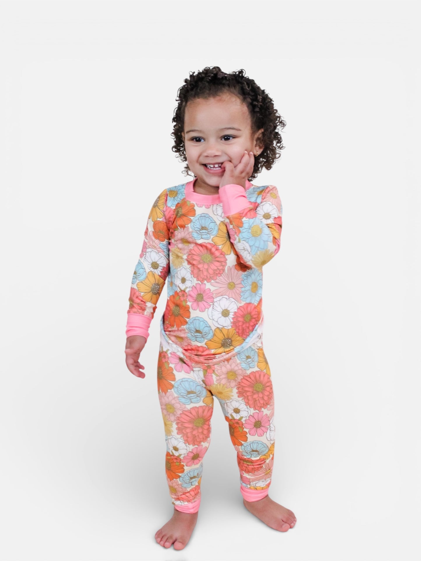 A young child with curly hair stands barefoot, smiling with one hand on their cheek, dressed in the Retro Bloom Jammies Set by Tiny Tot Bundles, featuring pink, orange, and blue floral patterns on a white background against a plain white backdrop.