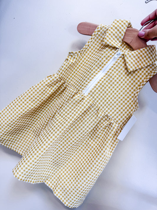The Buttercup Picnic Dress by Tiny Tot Bundles is a sleeveless yellow and white gingham dress with a collar and front white zipper, shown on a wooden hanger against a light background.
