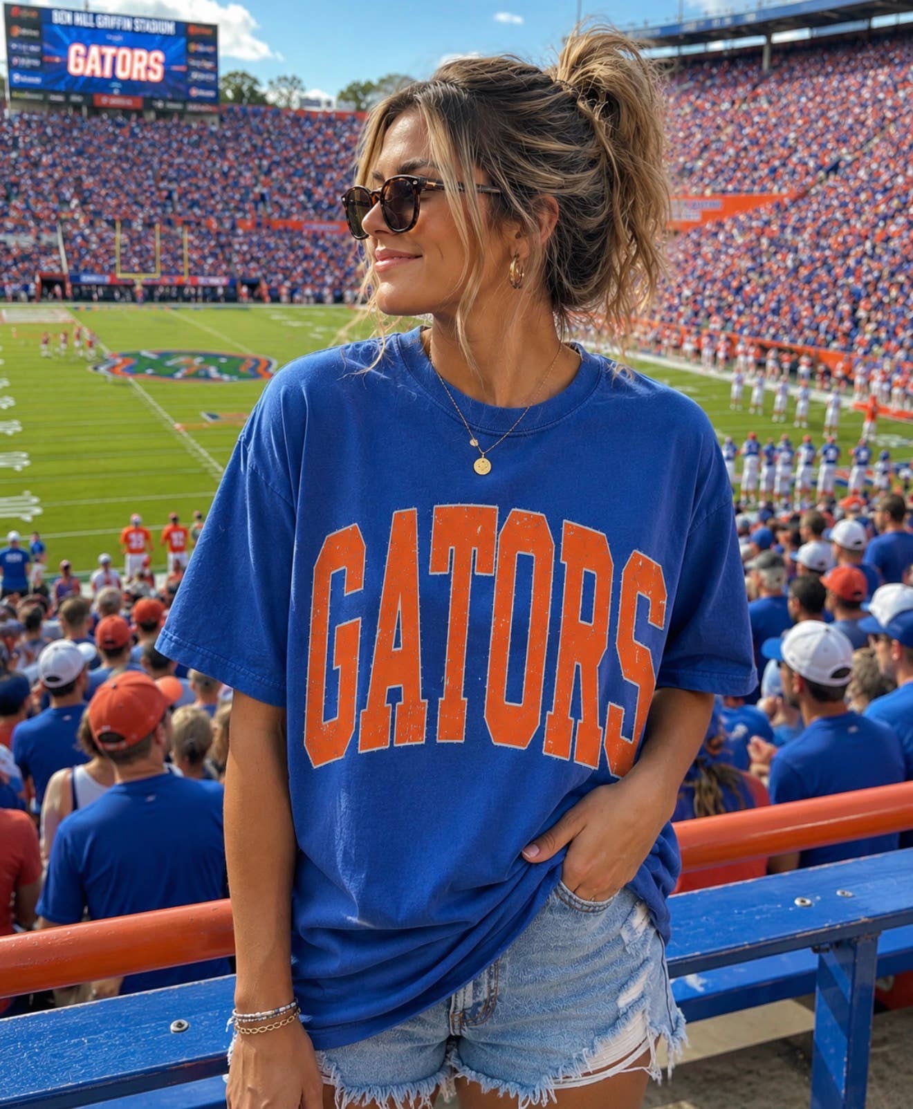 A woman wearing sunglasses, the oversized Gators Graphic Tee from Three Threads Branding Co., and denim shorts stands in a crowded football stadium filled with fans, with the field and scoreboard visible in the background.
