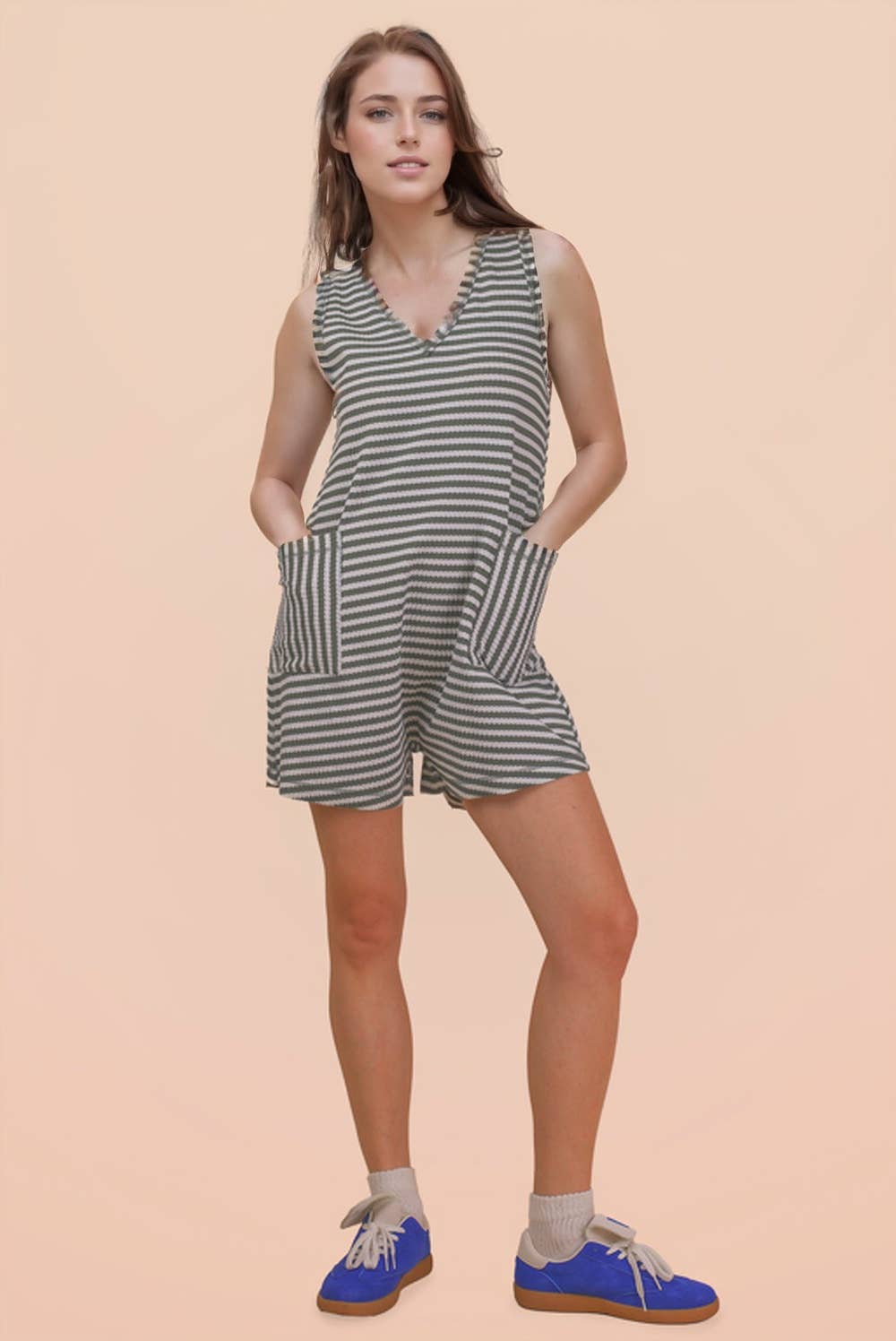 A woman stands against a light beige background, wearing the VERY J Striped Knit Casual Romper, which is sleeveless with black and white stripes and features large front pockets. She completes her look with beige socks and blue sneakers.