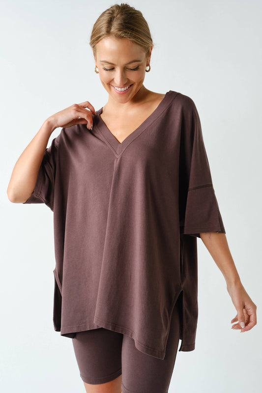 A woman smiles while wearing the Switchback Pima Cotton Set from Mono B, featuring a loose, brown V-neck top and matching brown bike shorts. She stands against a plain white background, gently holding the neckline of her shirt with one hand.