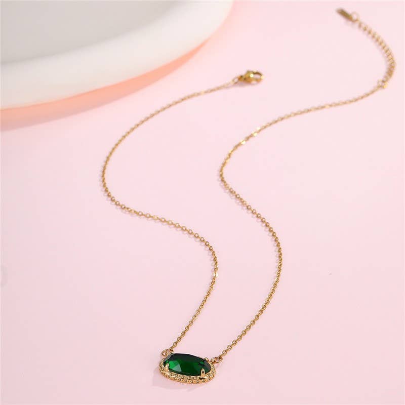The JOSSLYN by wall to wall Emerald Pendant Necklace, featuring a green oval gemstone set in delicate gold, rests on a light pink surface near a white dish.