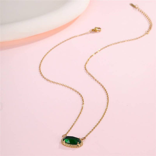 The JOSSLYN by wall to wall Emerald Pendant Necklace, featuring a green oval gemstone set in delicate gold, rests on a light pink surface near a white dish.