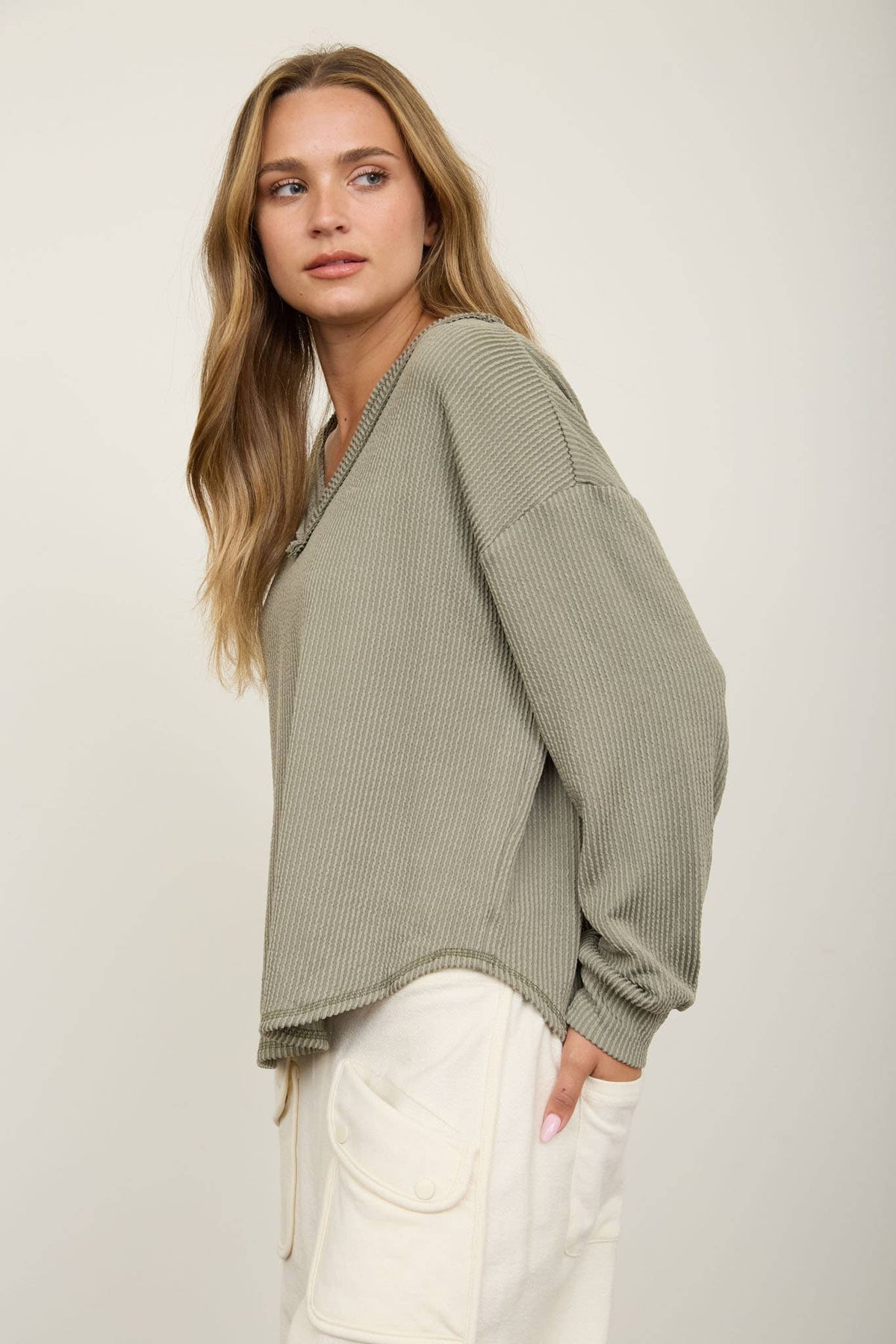 A woman with long, wavy hair models the Blu Pepper Olive V-Neck Long Sleeve Top, pairing the loose, ribbed sweater with cream pants featuring large pockets. She stands sideways against a plain background, glancing over her shoulder.