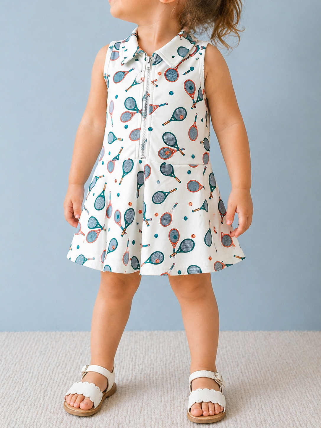 A toddler wears the Tiny Tot Bundles Green Quarter Zip Tennis Dress, featuring a tennis-themed print. She stands on a light carpet in white sandals against a light blue background, with her face partially out of view.