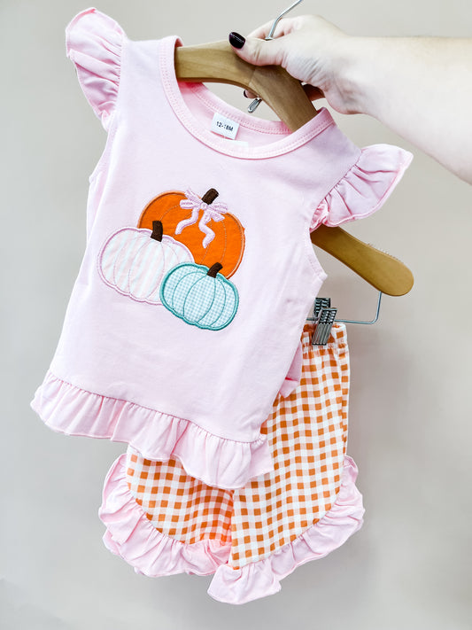 A hand holds the Tiny Tot Bundles “Sweet As Pumpkin Set,” a pink toddler outfit with ruffled sleeves and hem, featuring embroidered pumpkins on the top and orange gingham shorts, both displayed on wooden hangers.