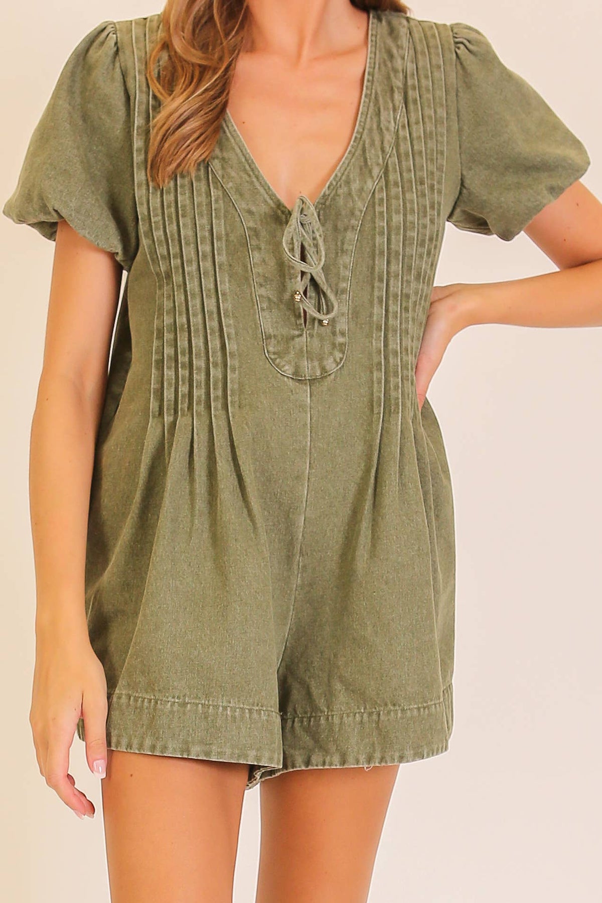 A woman wearing the Lumiere Desert Muse Utility Romper in olive green, featuring a pleated front, lace-up neckline, and puffed short sleeves.