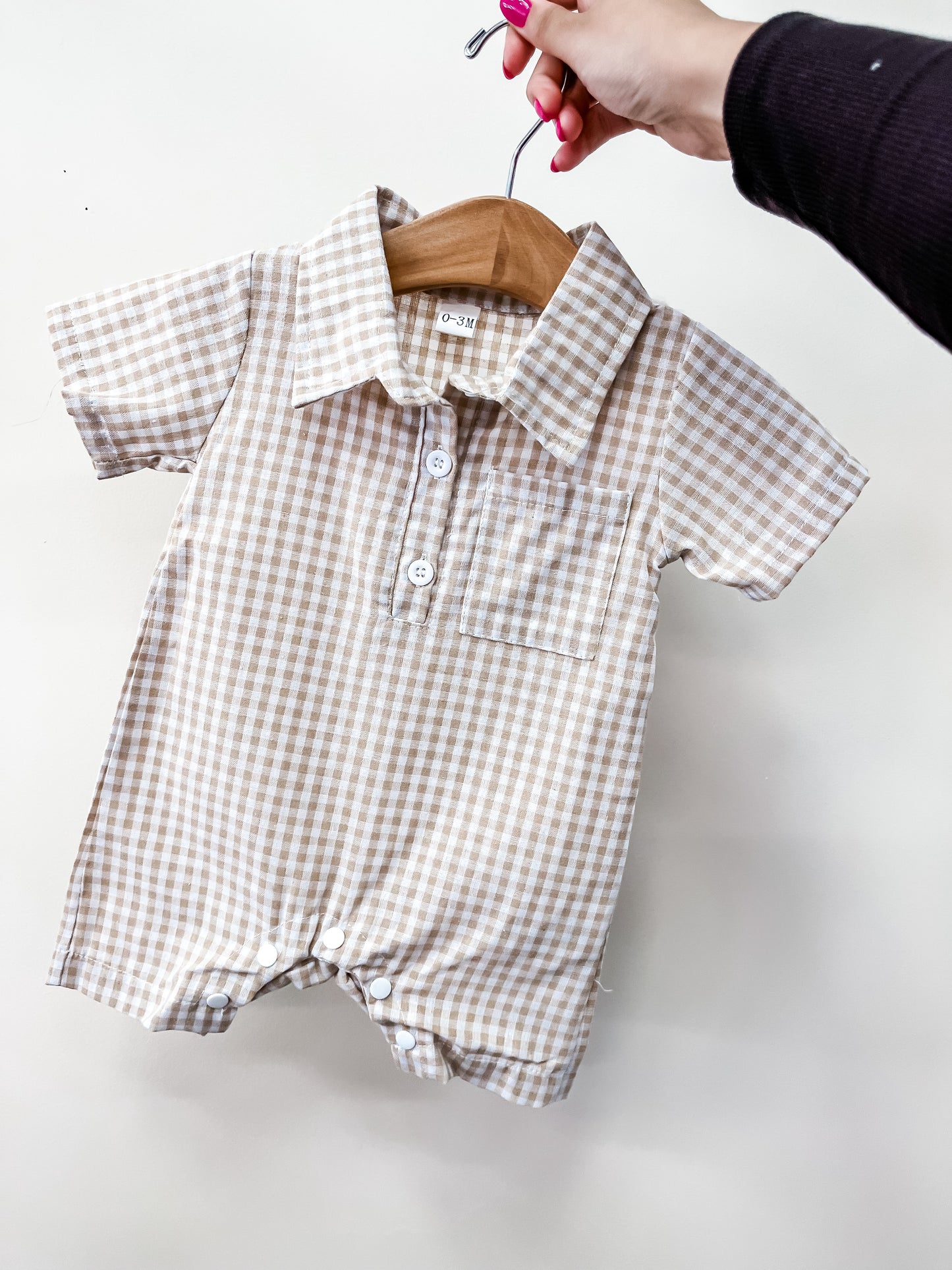 A hand holds the Tiny Tot Bundles Beige Gingham Collared Romper, a baby outfit with short sleeves, front buttons, collar, and a small chest pocket, displayed on a wooden hanger against a plain white background.