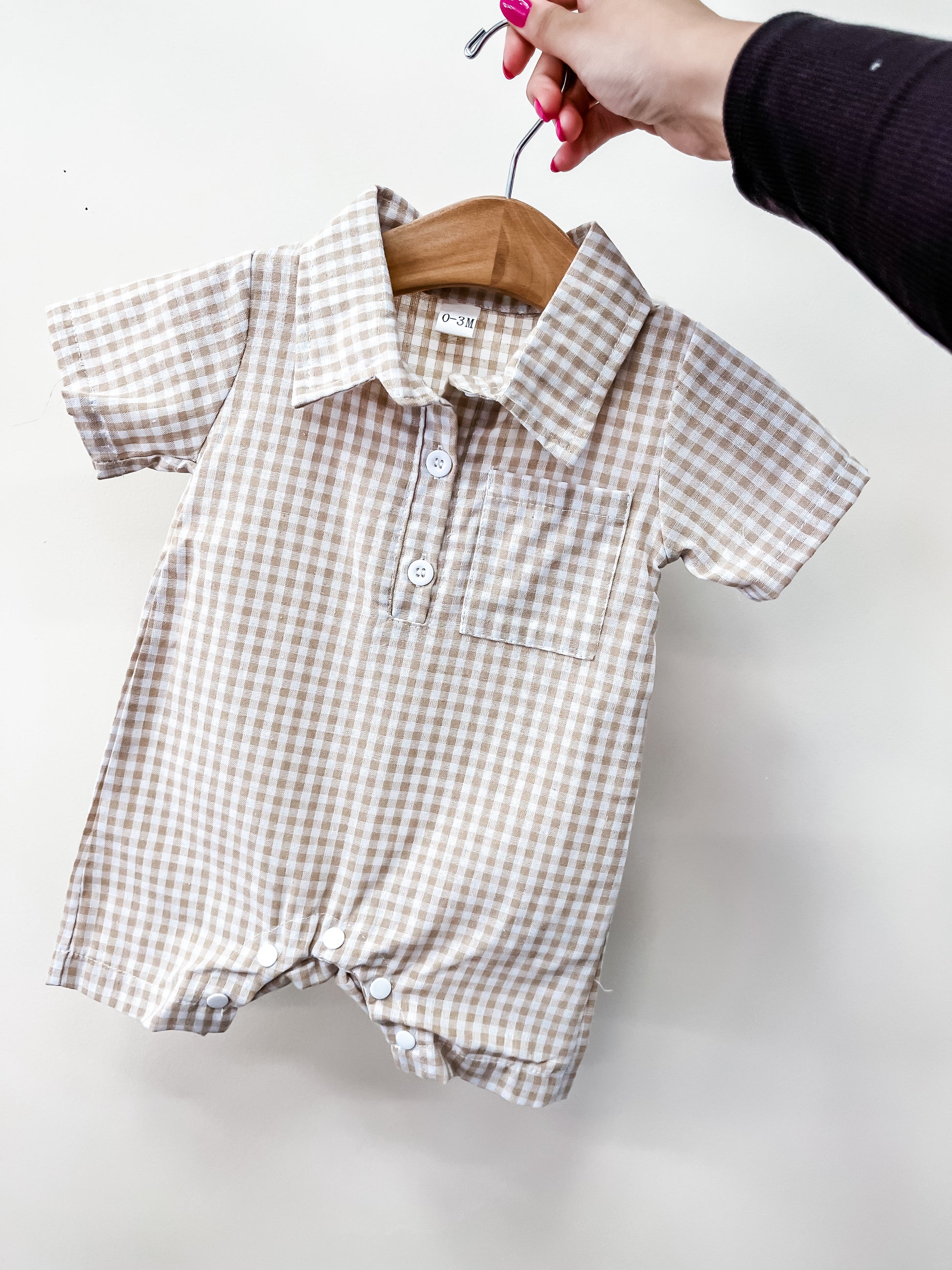 A hand holds the Tiny Tot Bundles Beige Gingham Collared Romper, a baby outfit with short sleeves, front buttons, collar, and a small chest pocket, displayed on a wooden hanger against a plain white background.