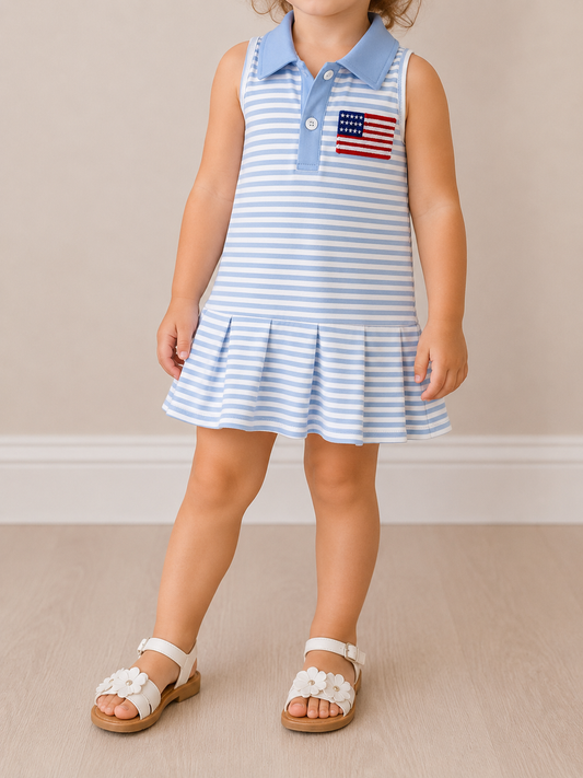 A young child wears the Tiny Tot Bundles Little Miss Americana Dress—a sleeveless light blue and white striped dress with a pleated skirt and American flag patch—paired with white flower sandals, standing on a light wood floor indoors.