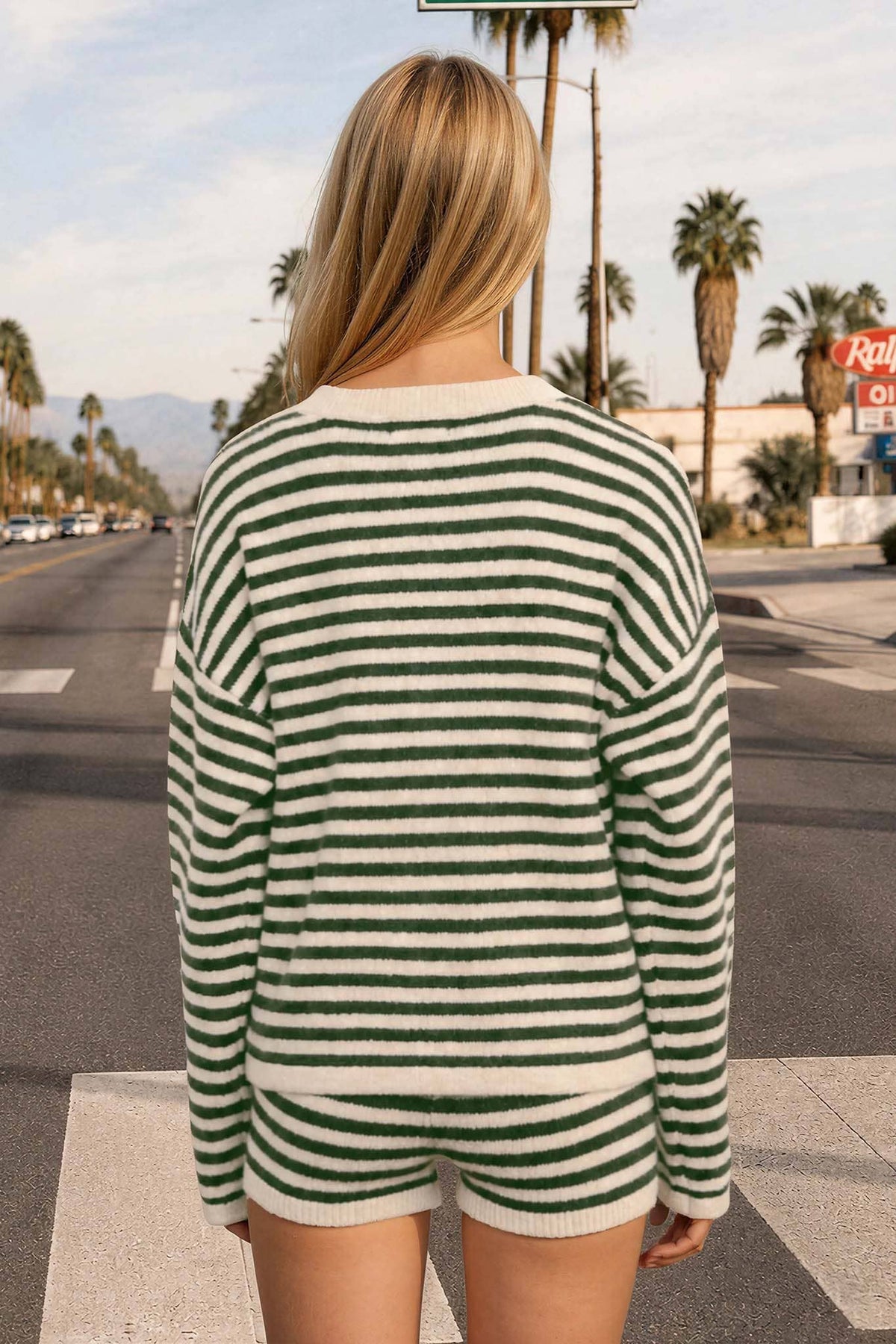 A person with long blonde hair stands at a crosswalk, facing away, wearing The Workshop Studio’s Green Stripe Long Sleeve Cardigan. Palm trees and a motel sign can be seen along the road.