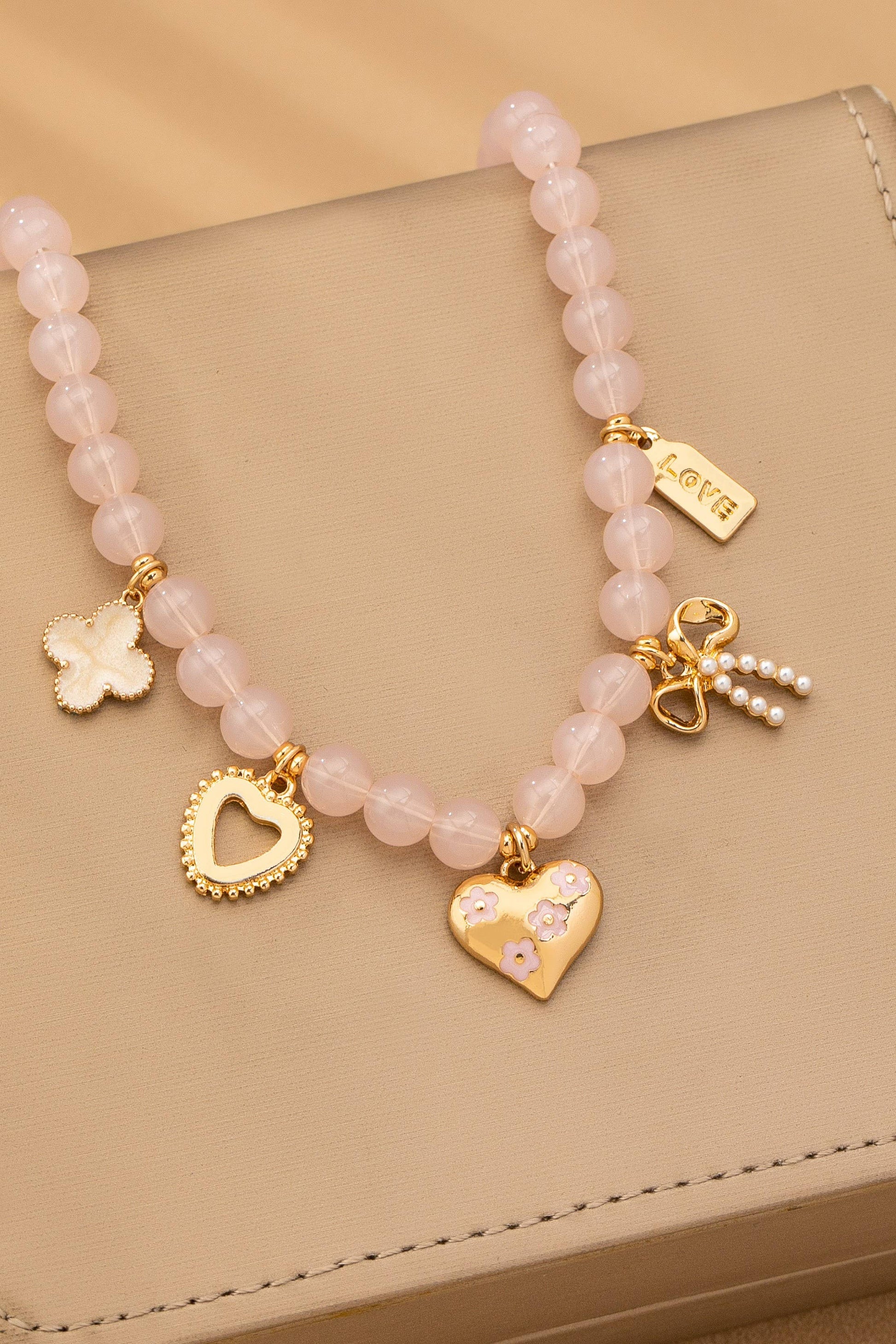 The JOSSLYN by wall to wall Beaded Charm Necklace features pink beads and gold and pearl charms—including a heart, flower, butterfly, bow, and “love” tag—elegantly displayed on a beige surface.