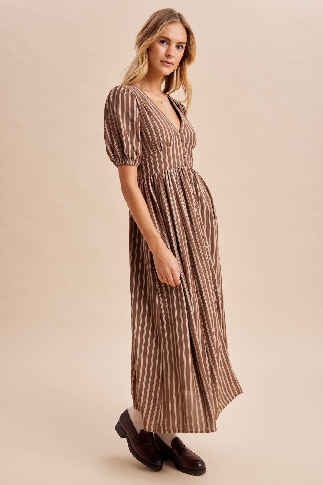 A woman wears an In Loom Stripe Button Down Maxi Dress, a brown and white vertically striped maxi with puff sleeves and a button-front closure, paired with dark loafers and white socks.
