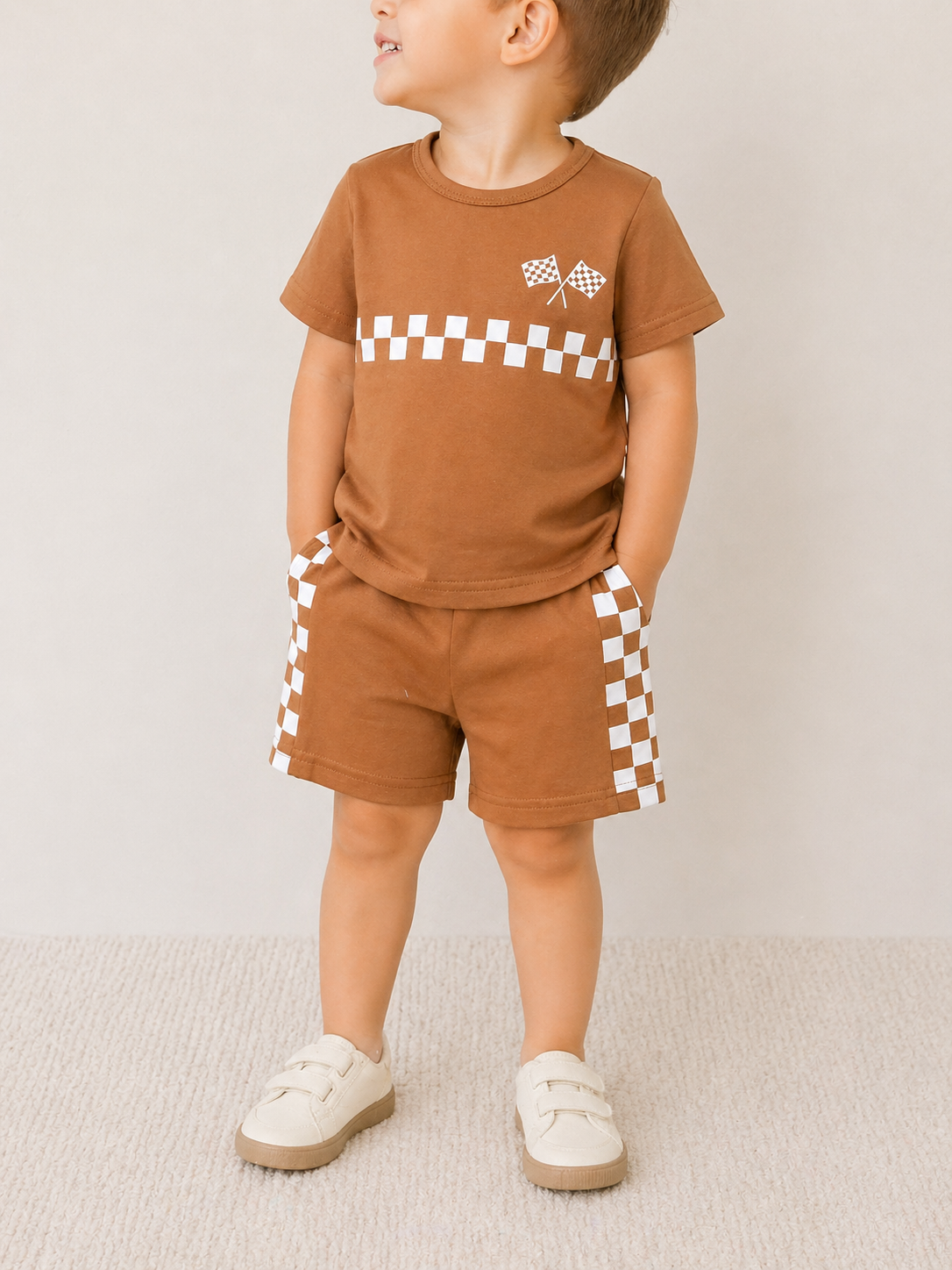 A young child models the Tiny Tot Bundles Rust Racer Check Set—brown t-shirt and white checkered shorts—paired with beige sneakers, standing against a light background with hands in pockets, looking to the side.
