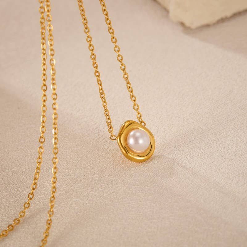 The Tarnish Free Pearl Necklace by JOSSLYN by wall to wall features a round white pearl pendant set in a wavy gold frame, elegantly displayed on a soft beige surface.