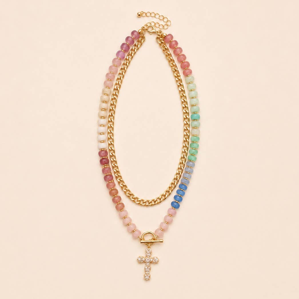 A JOSSLYN by wall to wall Multi Layer Pendant Resin Beaded Necklace featuring colorful beads, a gold chain, and a gold and rhinestone cross pendant is displayed on a light beige background.