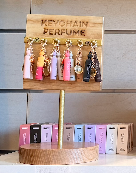 A wooden display stand holds Pureamor Perfume Oil Keychains, each featuring a gold keyring and vibrant colors. Boxes of Pureamor perfumes are lined up at the base, and a sign above reads “PERFUME OIL KEYCHAINS.”.