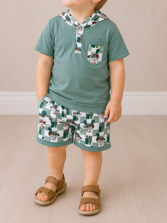 A young child stands indoors wearing the Pond Patrol Short Set by Tiny Tot Bundles, featuring a teal short-sleeve hoodie with a patterned pocket and matching shorts. The child's face is not visible.